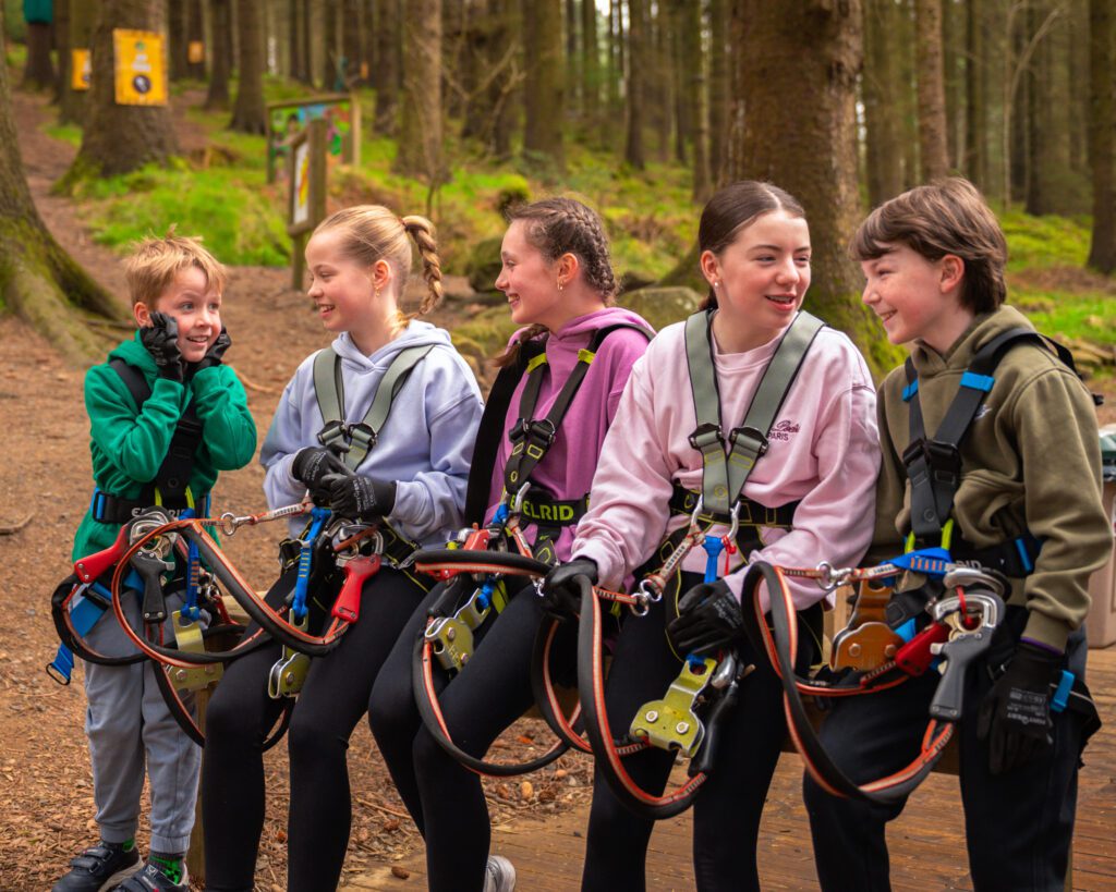 Five children in harnesses sit on a bench in a forest, preparing for a ziplining or ropes course activity, smiling and talking together.