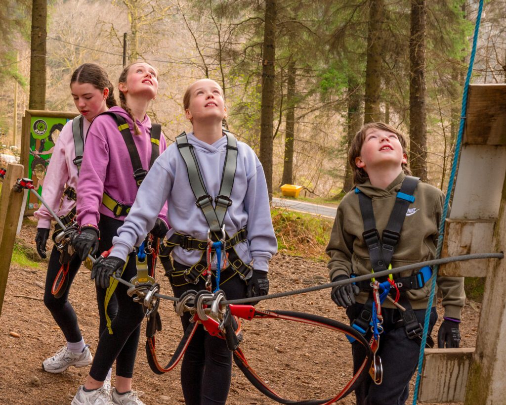 Four children wearing harnesses and gloves stand outdoors in a forest area, looking up at a ropes course or climbing structure.