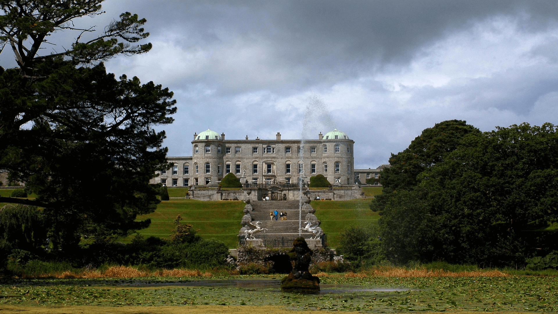 A large stone mansion with green domes sits atop a terraced hill, with statues lining the central staircase and a pond with a fountain in the foreground.
