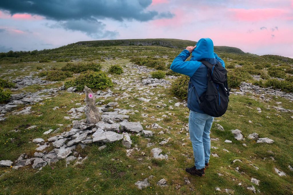 A person in a blue jacket and jeans stands on rocky terrain, looking towards a distant hill under a cloudy sky at sunset, capturing the spirit of Outdoor Activities Ireland.