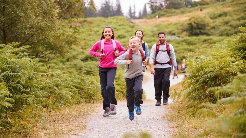 Two children run ahead while two adults walk behind them on a gravel trail through a green, wooded area. All are wearing backpacks and outdoor clothing, embracing Irish Family Adventures and encouraging kids off screens into nature.