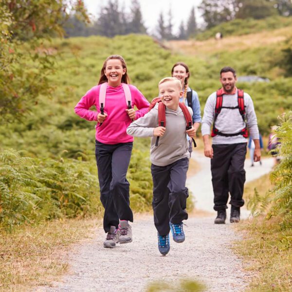 Two children run ahead while two adults walk behind them on a gravel trail through a green, wooded area. All are wearing backpacks and outdoor clothing, embracing Irish Family Adventures and encouraging kids off screens into nature.