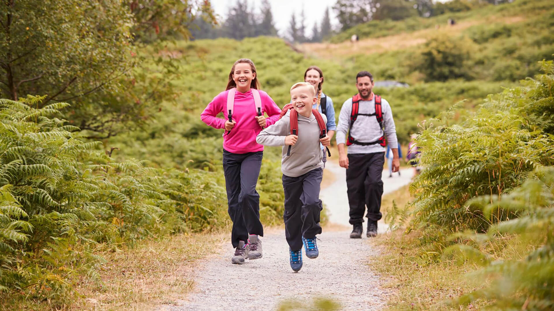 Two children run ahead while two adults walk behind them on a gravel trail through a green, wooded area. All are wearing backpacks and outdoor clothing, embracing Irish Family Adventures and encouraging kids off screens into nature.
