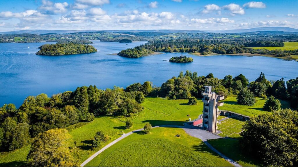 Aerial view of a lakeside park with a modern tower, trees, walking paths, grassy fields, and several forested islands—an ideal spot for Active Kids Parties or fresh Birthday Party Ideas Ireland under a partly cloudy sky.