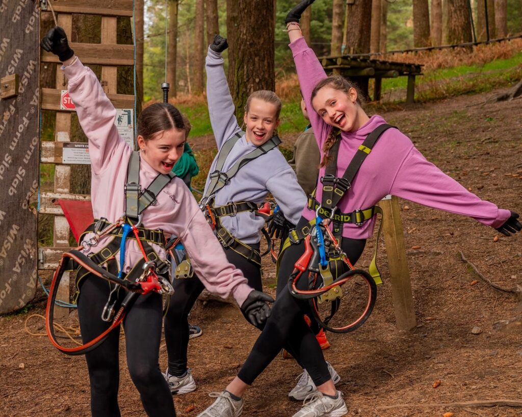 Three girls wearing safety harnesses pose and smile in a wooded outdoor adventure park—perfect for Active Kids Parties or unique Teens Birthday Ideas—standing on a dirt path near climbing equipment.