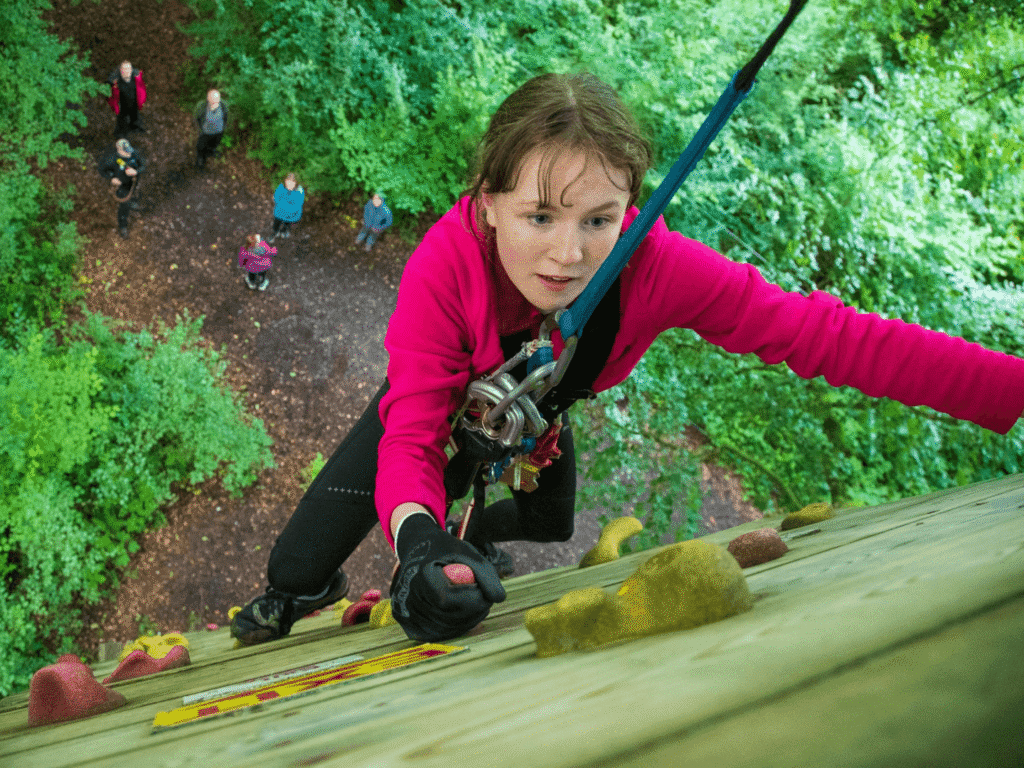 A person wearing a pink jacket and safety gear climbs an outdoor rock wall, demonstrating how adventure activities in Ireland can help overcome fear, while several people watch from the ground below in a wooded area.