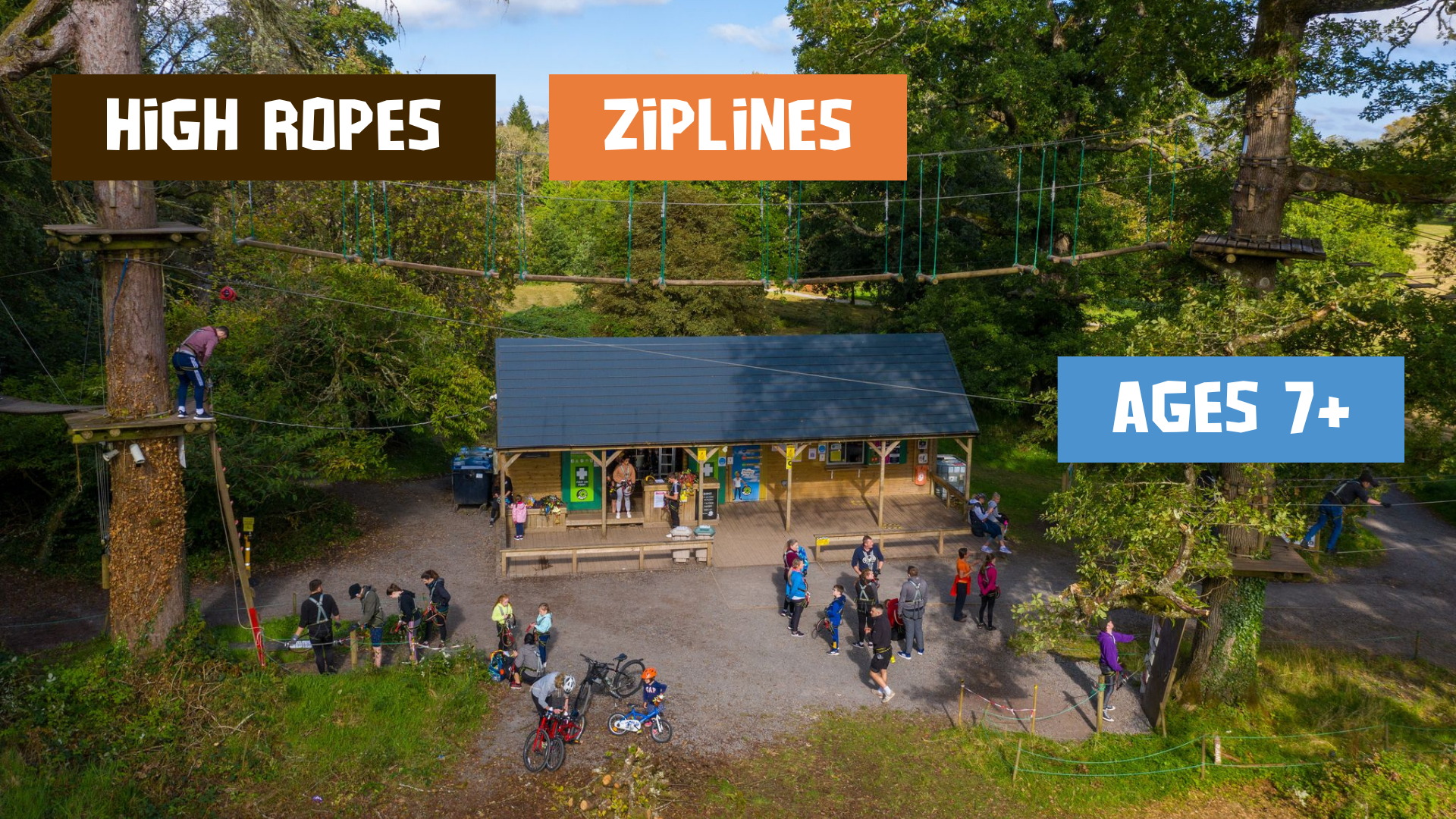 Aerial view of Kildare outdoor adventure park with people walking near a building; high ropes and zipwire courses winding through the trees; signs display activities and age requirement of 7 and upwards.