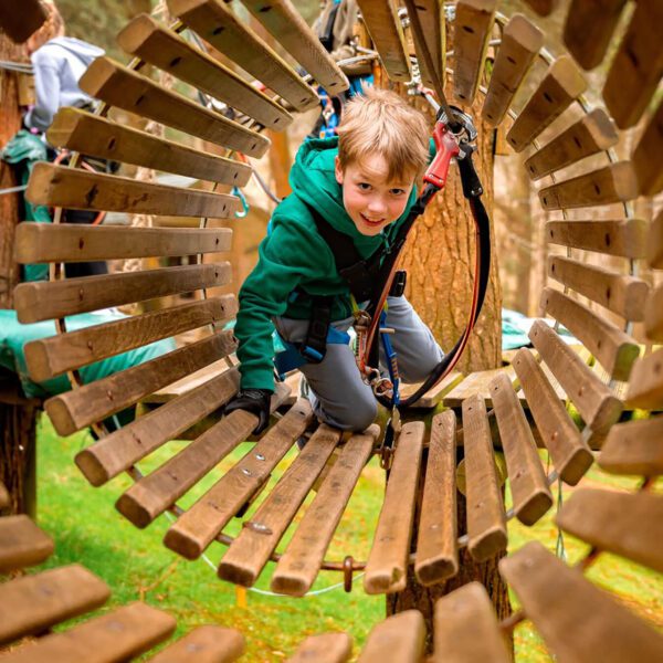 A young boy wearing a green jacket crawls through a wooden tunnel at an outdoor adventure park in Ireland, secured with a safety harness, as he takes part in adventure activities to build resilience and overcome fear.