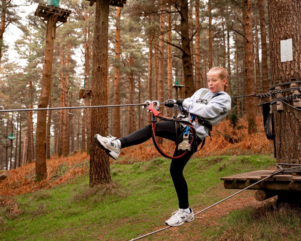 A young person in a safety harness prepares to zipline in a forest adventure park, balancing on a cable with one foot raised—an exciting way to build resilience and overcome fear through adventure activities in Ireland.