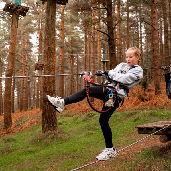 A young person in a safety harness prepares to zipline in a forest adventure park, balancing on a cable with one foot raised—an exciting way to build resilience and overcome fear through adventure activities in Ireland.