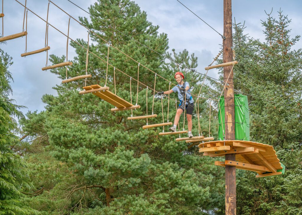 A person in a helmet and harness crosses a suspended rope bridge on a high ropes course at Zipit at Djouce Park, surrounded by tall trees.