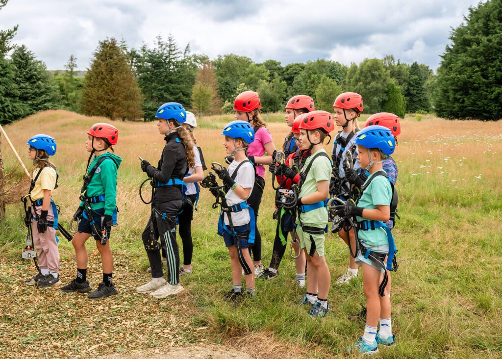 A group of children wearing helmets and harnesses stand outdoors in a grassy field at Zipit at Djouce Park, preparing for a climbing or ropes course activity.