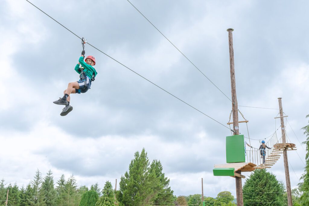 A person wearing a helmet and harness rides a zip wire at Zipit Forest Adventures, whilst another person waits on a wooden platform in the background.