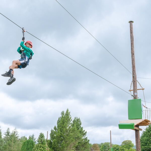 A person wearing a helmet and harness rides a zip wire at Zipit Forest Adventures, whilst another person waits on a wooden platform in the background.