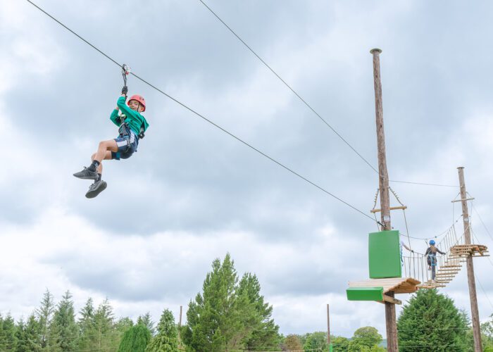 A person wearing a helmet and harness rides a zip wire at Zipit Forest Adventures, whilst another person waits on a wooden platform in the background.